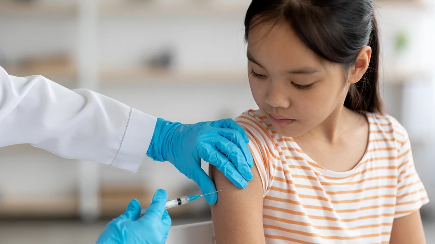 Unrecognizable doctor hands in protective gloves making vaccination against coronavirus of flu for little asian girl, home interior, closeup shot. Kids immunization against various disease concept